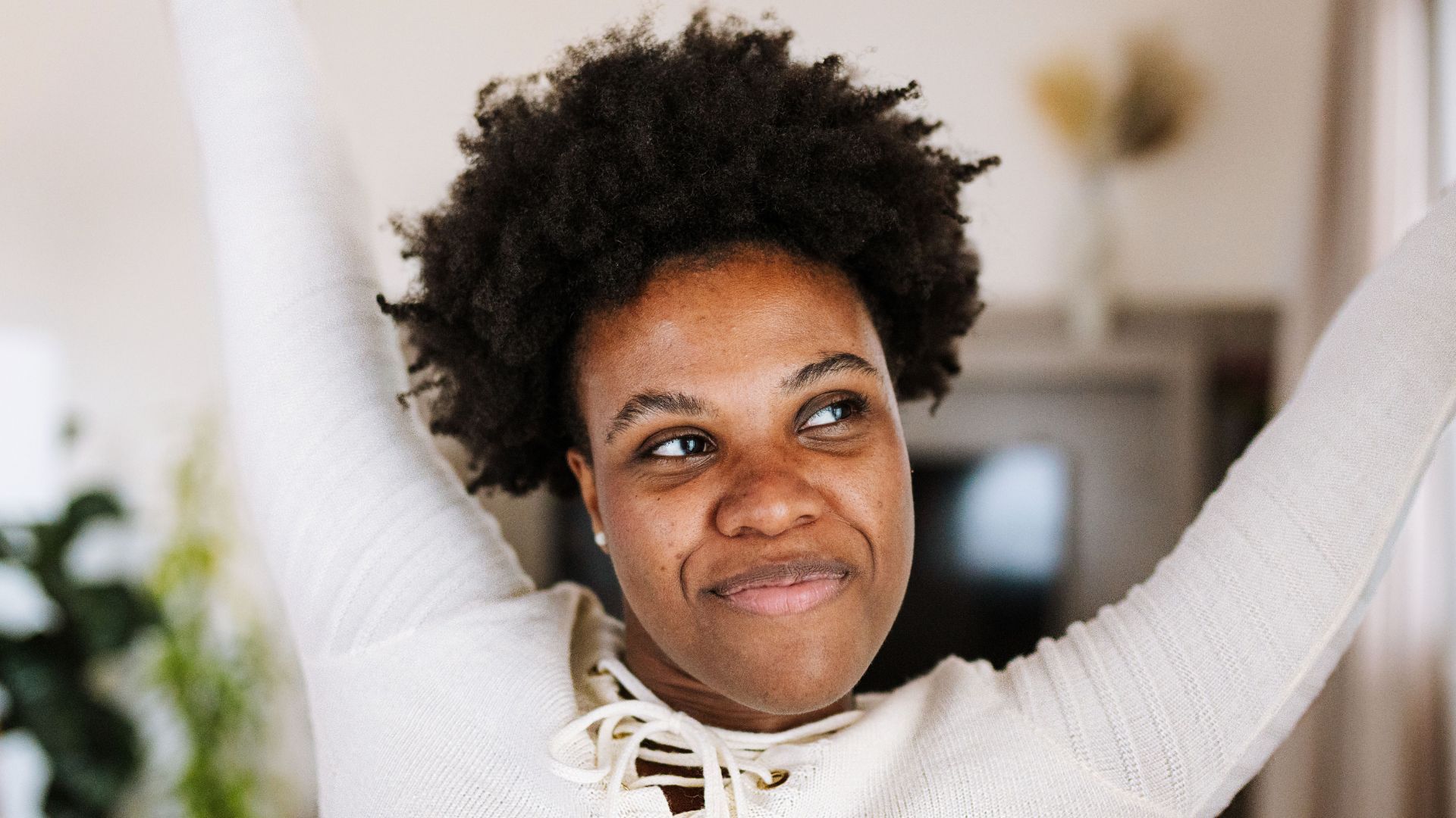 Woman with arms raised, smiling softly indoors.